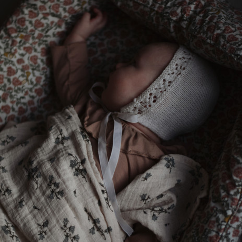 Baby liegt im Babynestchen und ist zugedeckt mit Musselin Swaddle in Beige mit Blumenmuster von Garbo & Friends.