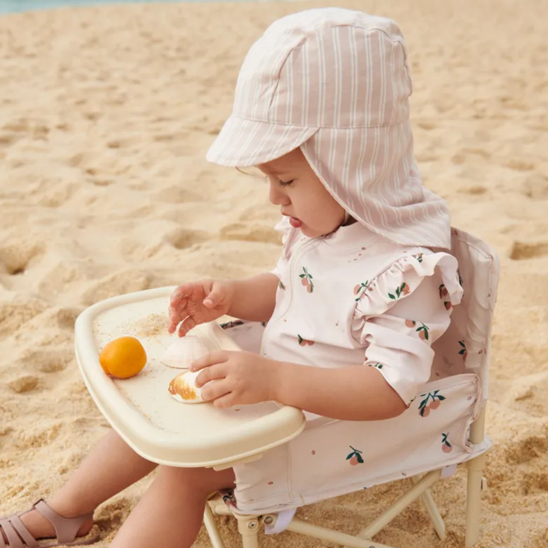 Baby sitzt am Strand in einem Strandstuhl und trägt einen rosa Sonnenhut und den Badeeinteiler für Babys in Beige mit Pfirsichmuster von Liewood. 
