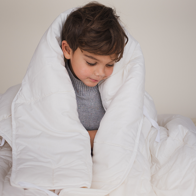 Junge sitzt und hat die Bettdecke für Kinder von Murmunto in Weiss um sich gehüllt. 