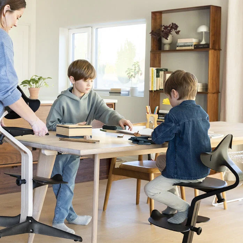 Junge sitzt am Tisch auf Nomi Hochstuhl für Kinder in Schwarz von Stokke. Ein anderer Hochstuhl wird von der Mutter an den Tisch getragen und der zweite Junge spielt im Stehend am Tisch.