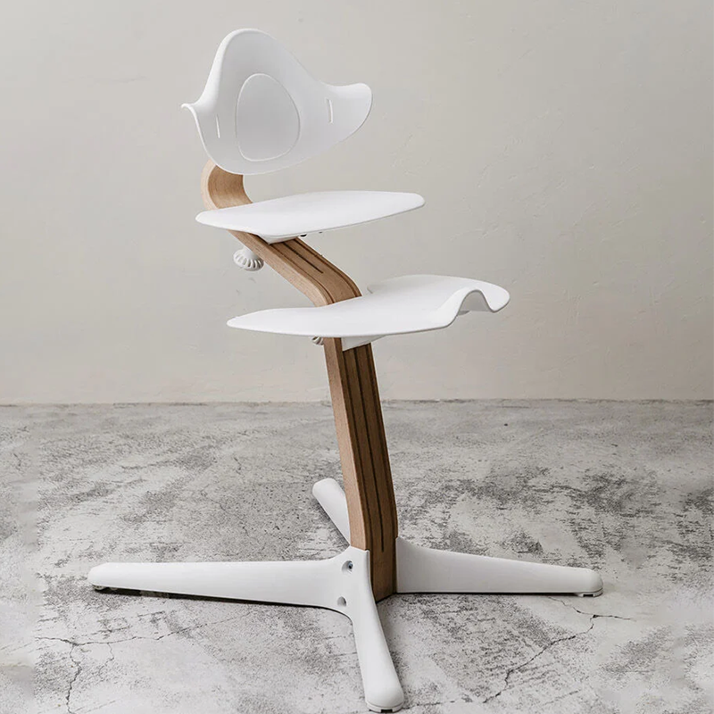 Nomi Hochstuhl mit Mittelbalken aus Walnussholz und Rückenlehne, Sitz, Fussauflage und Füsse in Weiss von Stokke steht auf Betonboden. 
