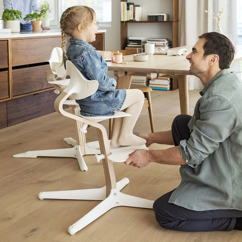 Mädchen sitzt auf dem Nomi Hochstuhl mit Mittelbalken aus Holz und Sitz, Rückenlehne, und Füsse in Weiss von Stokke und vor dem Stuhl kniet der Vater. 