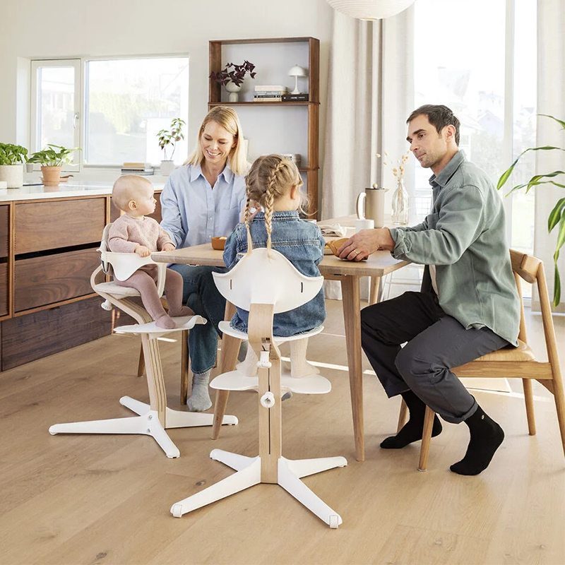 Familie sitzt am Tisch die Kinder sitzen auf dem Nomi Hochstuhl mit Mittelbalken aus Holz und Sitz, Rückenlehne, und Füsse in Weiss von Stokke. 