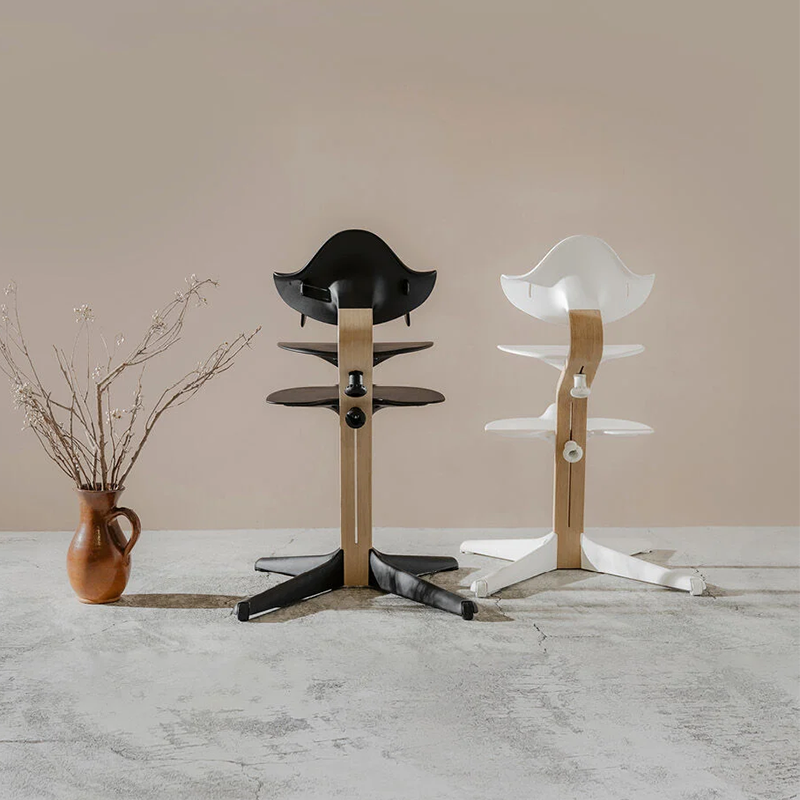 Nomi Hochstuhl mit Mittelbalken aus Holz mit Sitz, Rückenlehne, und Füsse in Weiss von Stokke steht neben Nomi Hochstuhl mit Mittelbalken aus Holz mit Sitz, Rückenlehne und Füsse in Schwarz von Stokke. 