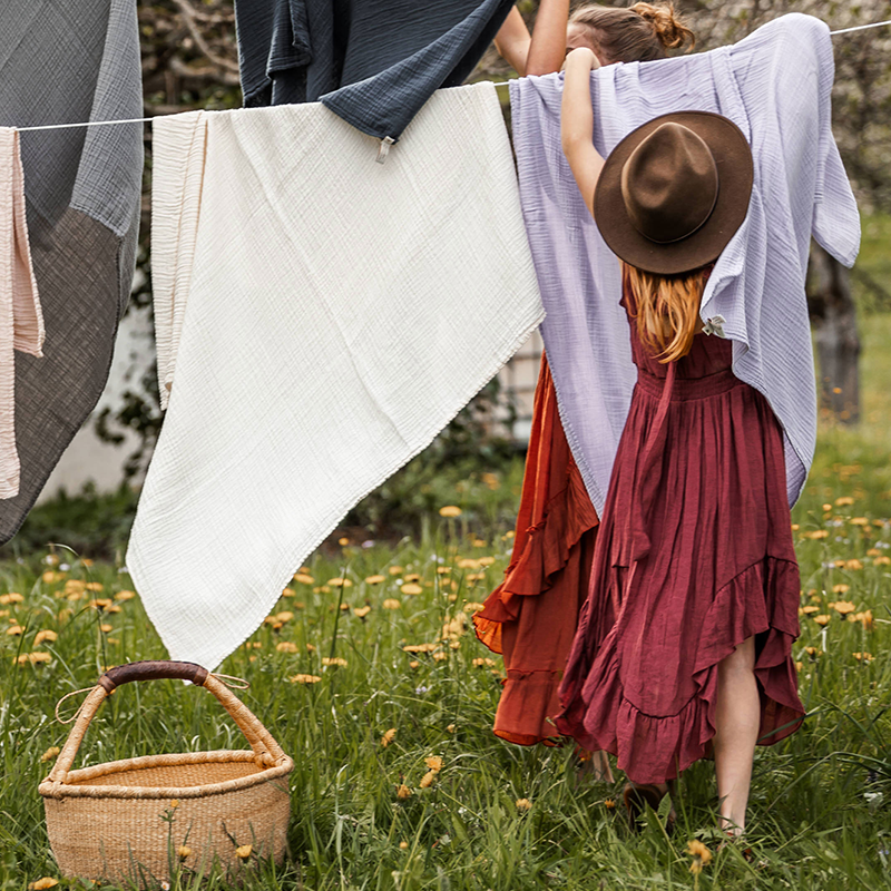 Frauen hängen Musselin Nooshie Decke in lila, beige, blau, grau und weiss von Petit Stellou auf. 