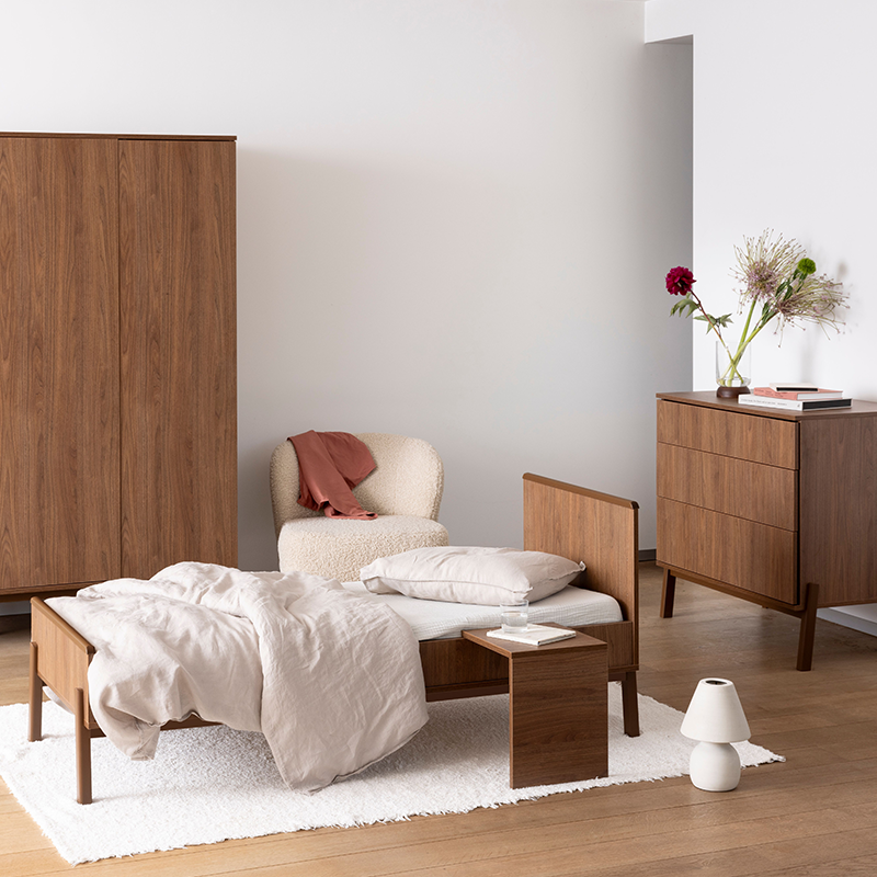 Juniorbett in Kastanienbraun mit Nachttisch mit weissem Kissen und weisser Decke steht auf einem weissen Teppich in einem Kinderzimmer. Im Hintergrund steht passender Kleiderschrank und passende Kommode.