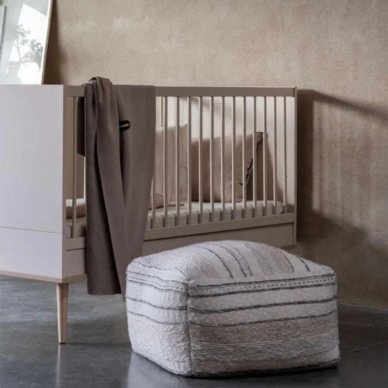 Babybett in Beige mit 4 Beinen aus massiver Buche in Babyzimmer. Pouf steht neben dem Babybett.