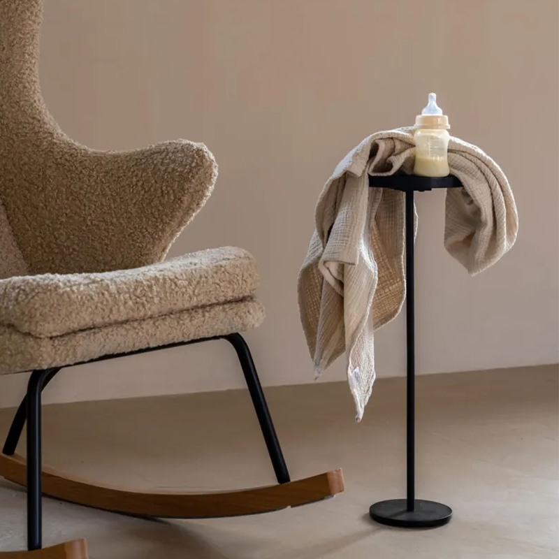 Beistelltisch mit runder Tischplatte in Schwarz von Quax neben einem Schaukelstuhl. Auf dem Tisch befindet sich ein Babyfläschchen und ein Spucktuch.