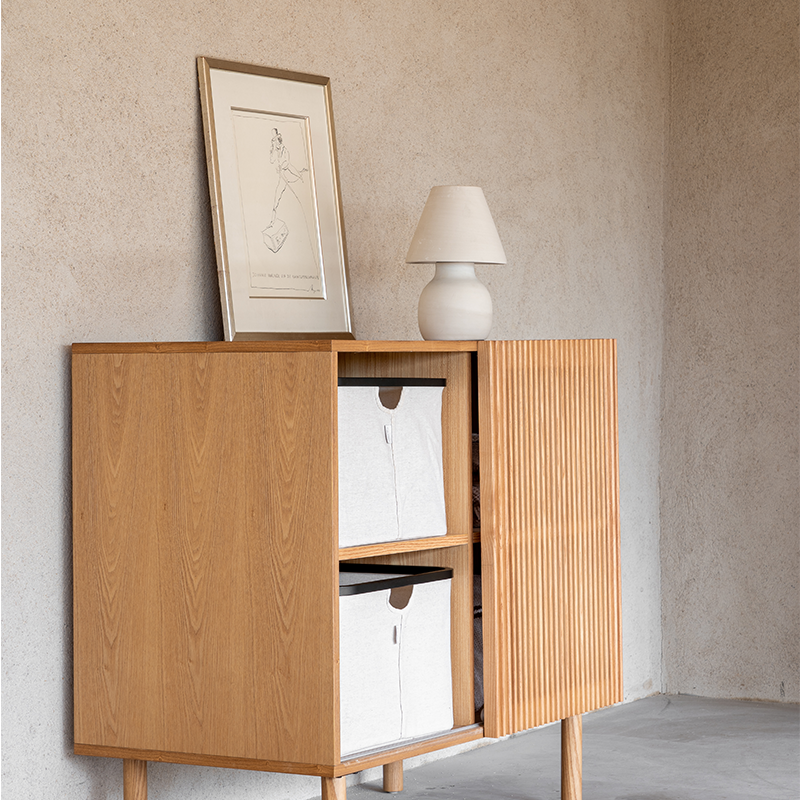 Kommode aus Ahornholz mit vier Ablagefächern und elegante Schiebetür mit Gitterstäben von Quax. Auf 2 Ablagefächern stehen Aufbewahrungskörbe und auf der Kommode steht ein Bild und eine Stehlampe.