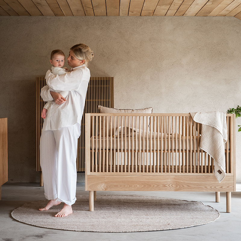 Mama hält Baby auf dem Arm und steht neben Babybett in Eschenoptik von Quax.