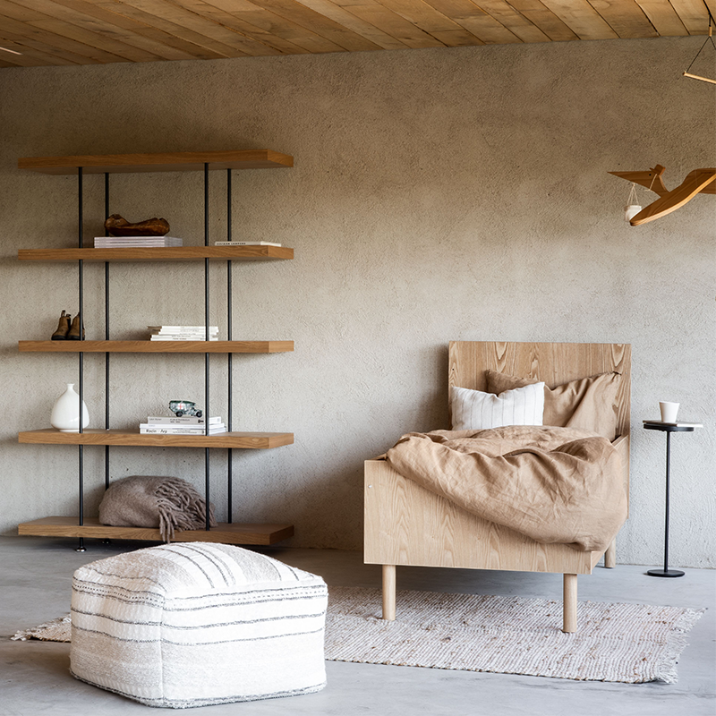 Juniorbett aus Eiche steht in einem Kinderzimmer. Neben dem Bett steht ein Bücherregal und ein kleines Nachttisch und vor dem Bett steht ein weisser Pouf.
