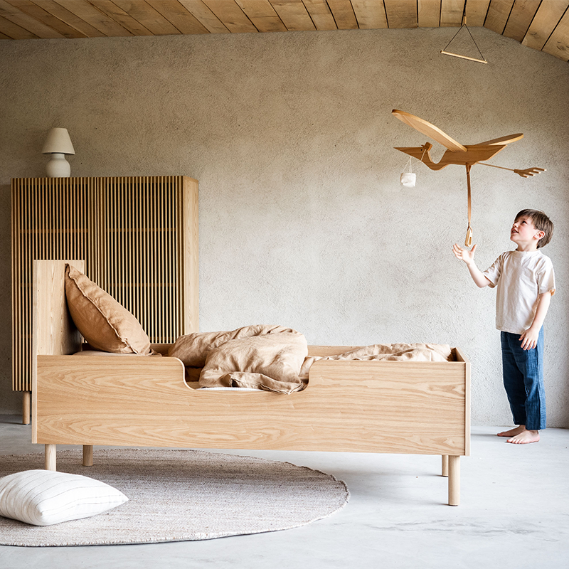 Juniorbett in Eichenoptik von Quax steht in Kinderzimmer. Ein Junge spielt mit einem Holzmobile, das an der Decke hängt.