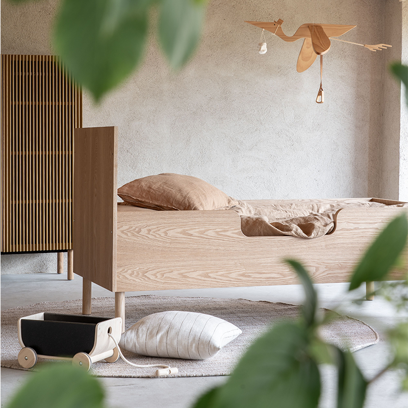 Juniorbett aus Eiche mit Bettwäsche in Beige steht in einem Kinderzimmer. Über dem Bett hängt ein Vogel aus Holz.