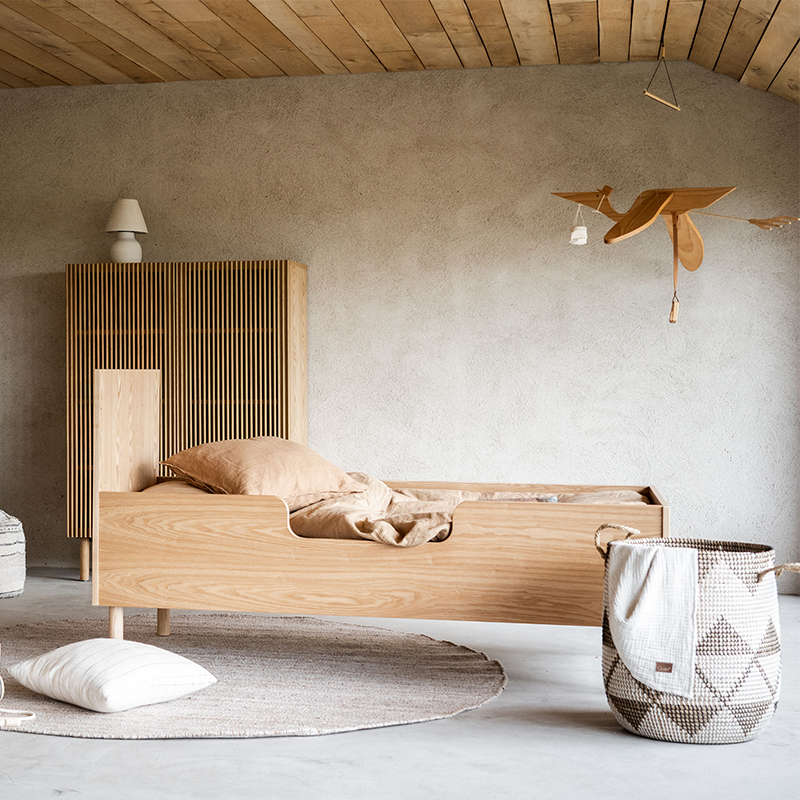 Juniorbett aus Eiche steht in einem Kinderzimmer. Hinter dem Bett steht ein Schrank aus Eiche und vor dem Bett ein Wäschekorb.