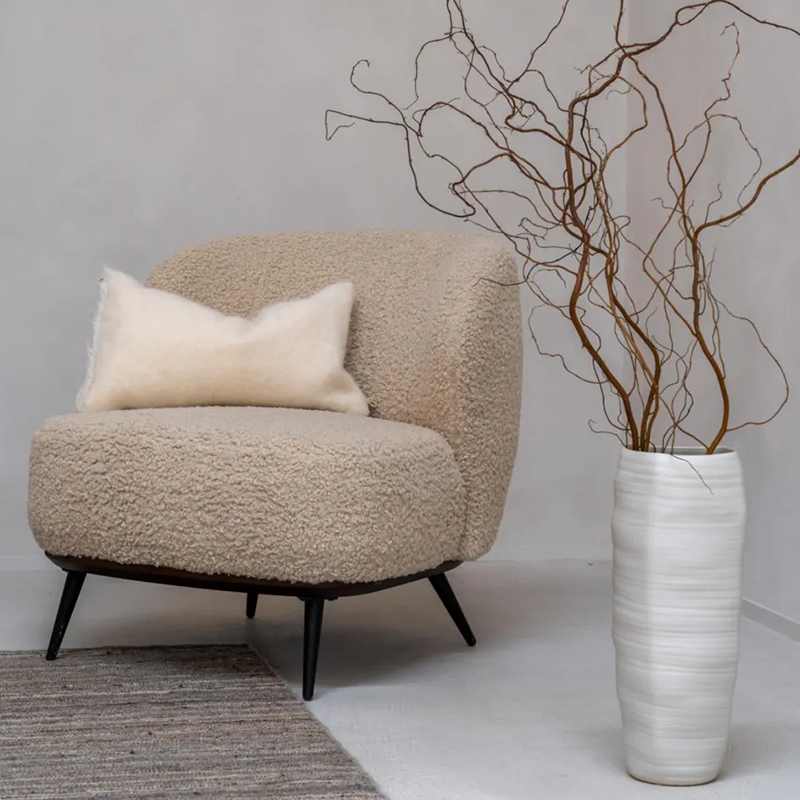 Loungesessel in Beige mit weichem Teddy-Bezug von Quax steht in einer Ecke neben einer grossen Vase mit Ästen drin. 
