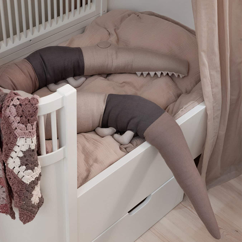 Kissen in Form eines Krokodils in Beigetönen von Sebra liegt in einem Babybett.