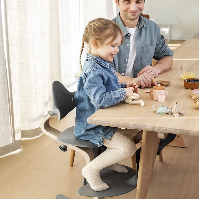 Mädchen sitzt am Tisch auf dem Nomi Hochstuhl für Kinder mit Mittelbalken aus Holz und Sitz, Rückenlehne  und Fussauflage und Füsse in grauem Plastik von Stokke und spielt mit seinem Vater. 