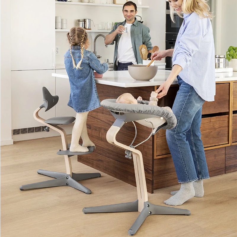 Mädchen ist auf dem Nomi Hochstuhl für Kinder mit Mittelbalken aus Holz und Sitz, Rückenlehne  und Fussauflage und Füsse in grauem Plastik von Stokke an der Kücheninsel Baby liegt im Nomi Hochstuhl in Grau und wird gefüttert. 
