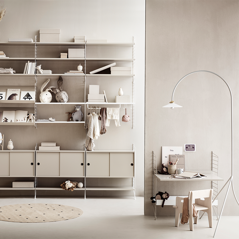 String Furniture Regalkombination mit Spielsachen und Kinderbüchern auf der linken Seite und Bodenregal in Beige mit einem Regalboden unten und mit einer Schreibtischplatte in der Mitte von String Furniture auf der rechten Seite. 