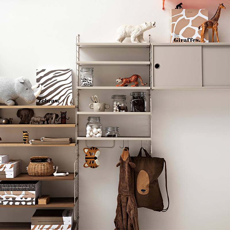 String Furniture Regalsytem in Beige in Kinderzimmer mit verschiedenen Tieren und Bücher und eingehängten Haken an denen Taschen und Kleidung hängt. 