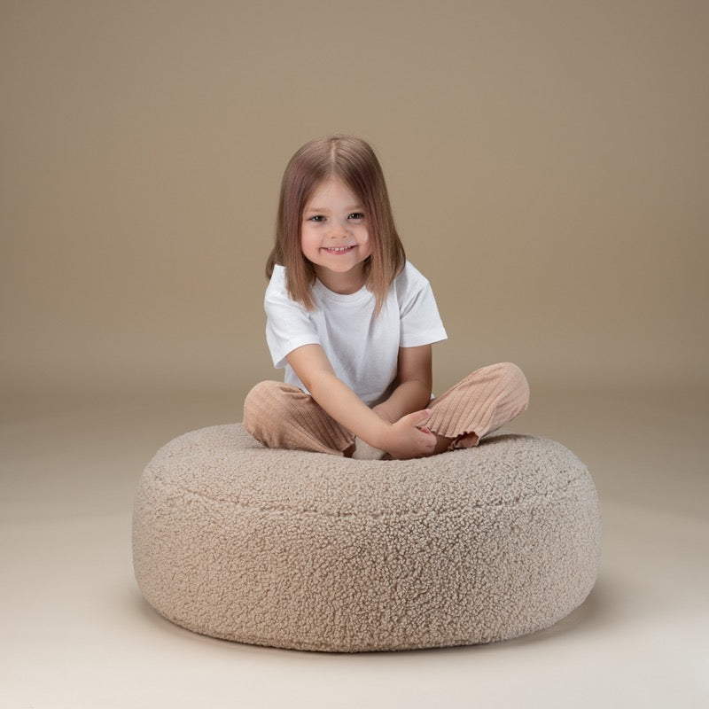 Kind sitzt im Schneidersitz auf dem runden Teddy-Ottoman in Biscuit-Beige von Wigiwama
