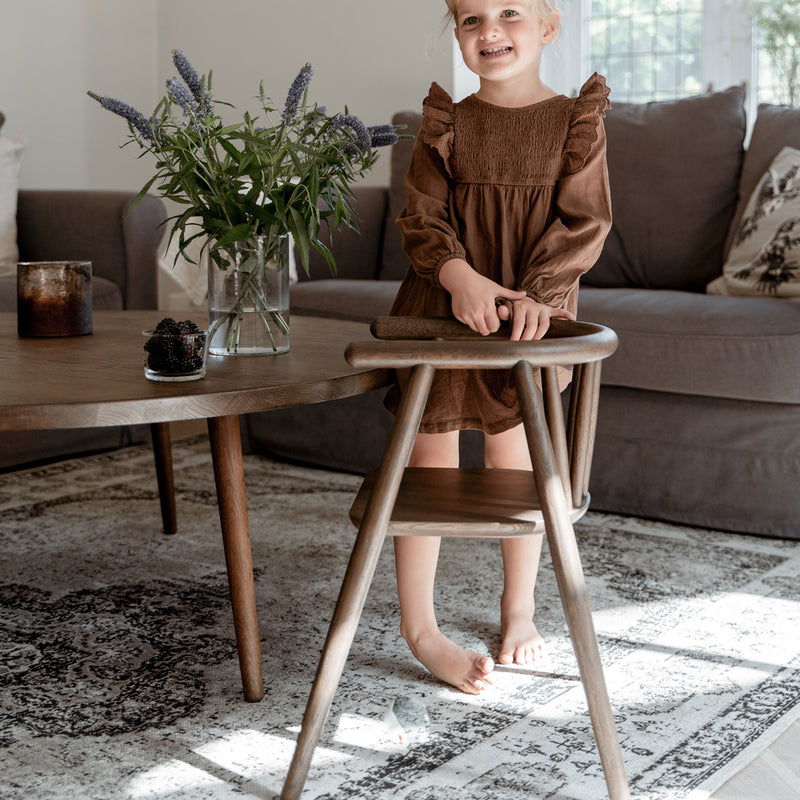 Mädchen steht am Tisch neben dem Oaklings Kinderstuhl 'STORM' in Smoked Oak. 